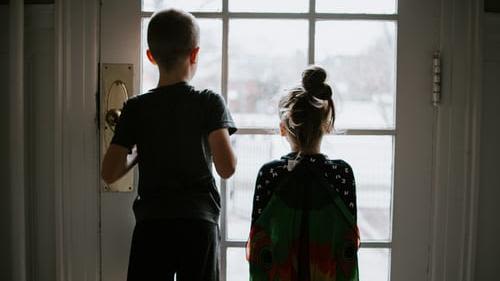 A taller boy and a girl with their backs turned looking out a glass paned door.