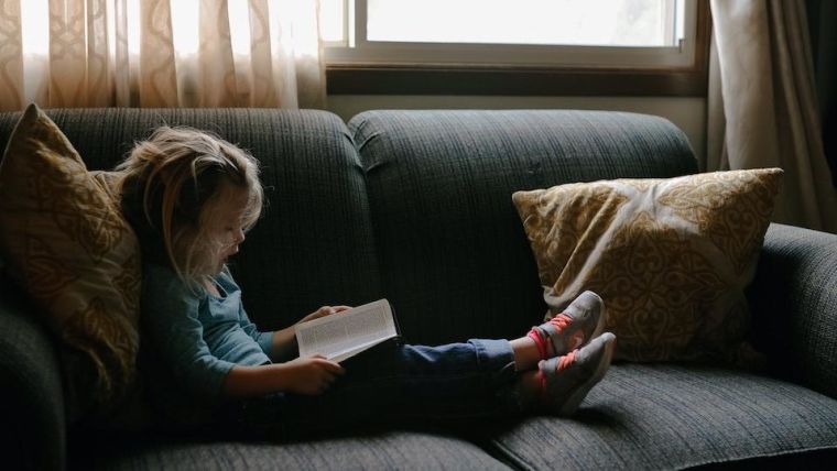 Young girl sitting on grey sofa reading a book.