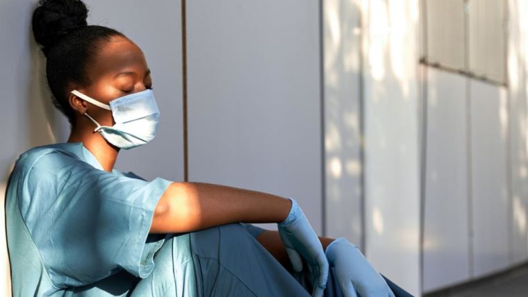 Female healthcare worker wearing a face mask sitting against a wall looking exhausted